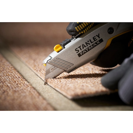 Photo of a STANLEY 0-11-800 utility knife cutting brown carpet, close-up on blade and gloved hand.