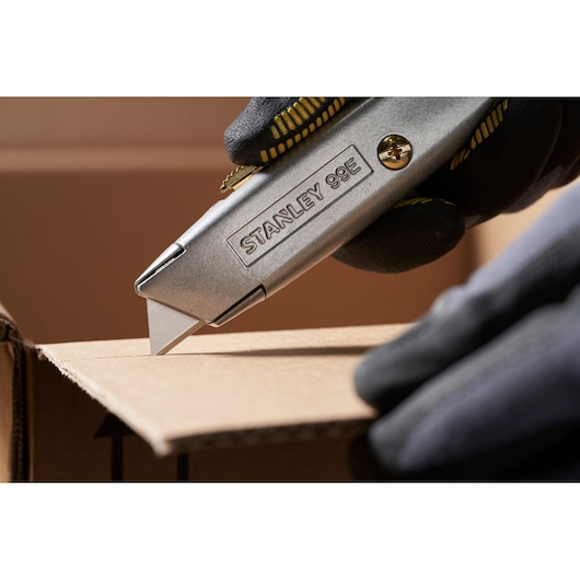 STANLEY 0-11-911 utility knife cutting cardboard, close-up side view, worker wearing black gloves.