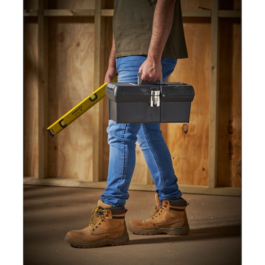 STANLEY 1-94-857 black toolbox and yellow level, carried by a person in work boots and jeans on construction site.