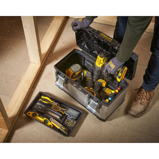 A person wearing gloves opens a Stanley toolbox containing various power tools and accessories. A removable tray with hand tools, screws, and a level is placed on the floor next to the box in a workshop setting.