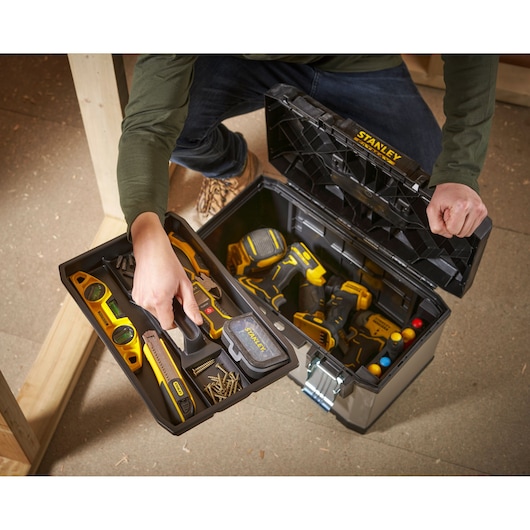 A person is opening a Stanley toolbox, which contains various hand tools and power tools, including a spirit level, screwdriver, wrench, screws, and drill. The tools are organized in a removable tray and in the lower compartment of the toolbox.