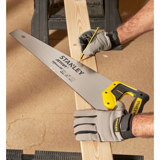 Stanley hand saw with yellow handle held on wood plank. Gloved hands mark the plank with a pencil for cutting.