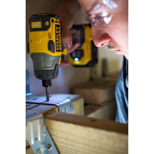 Photo of a person using a STANLEY FMC647D2 cordless impact driver on wood with metal brackets, close-up action shot.