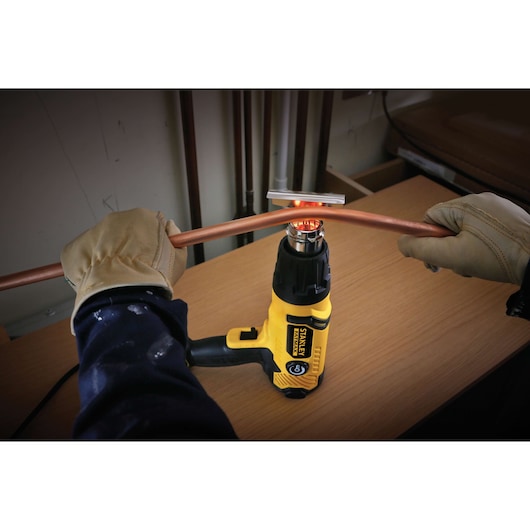 STANLEY FME670K heat gun in use bending a copper pipe, person wearing gloves holds pipe above tool on a workbench.