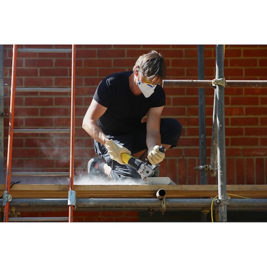 STANLEY FME811K corded angle grinder in use on construction site, worker wearing gloves and mask, brick wall background.