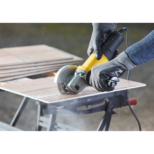 STANLEY FMEG210K angle grinder cutting tile, close-up view, yellow and black tool, held by gloved hands.