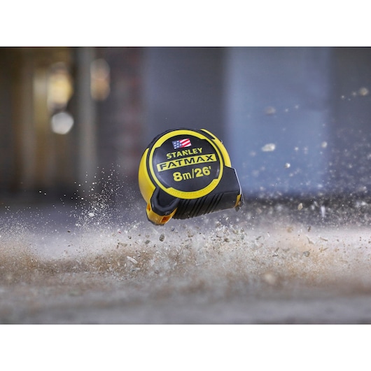 Stanley FMHT0-36326 FatMax tape measure 8m/26ft, yellow and black, in mid-air above dusty ground.