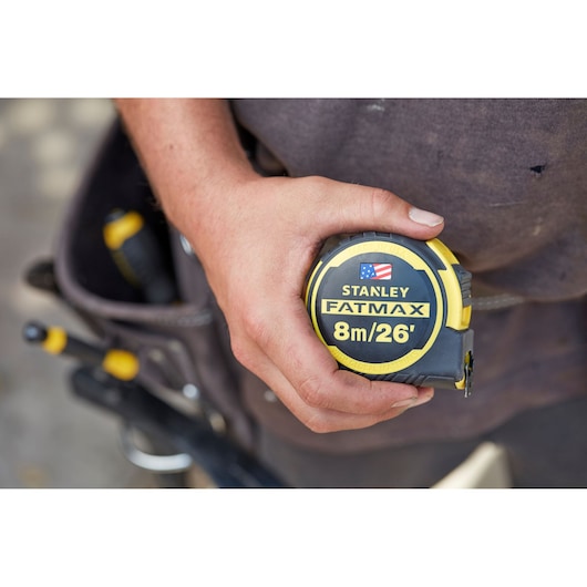 Photo of STANLEY FMHT0-36326 FATMAX tape measure, 8m/26', held in hand, tool belt in background.