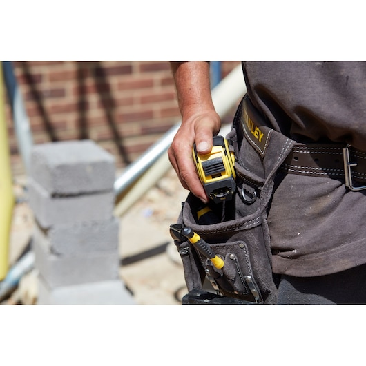 Photo of STANLEY FMHT0-36326 tape measure in tool belt on person's waist, construction site background.