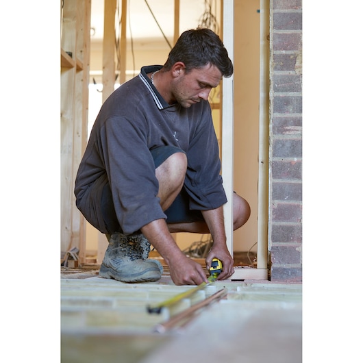 STANLEY FMHT0-36336 tape measure in use measuring floor in construction site, handheld by person squatting.