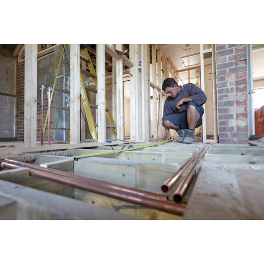 STANLEY FMHT0-36337 tape measure used on construction site by worker, shown with wooden framing and pipes.