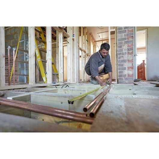 Photo of STANLEY FMHT0-36337 tape measure used on construction site over wooden floor joists and copper pipes.