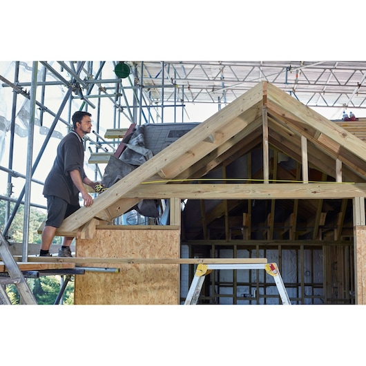 STANLEY FMHT0-36337 tape measure on roof construction, worker measuring wooden framework, outdoor site.