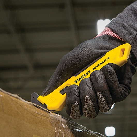 Stanley FMHT10358 utility knife with yellow handle, used to cut cardboard, held by gloved hand.