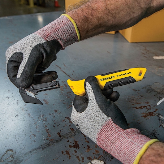 Stanley FMHT10358 utility knife held in gloved hands, yellow handle, blade compartment open, industrial setting.