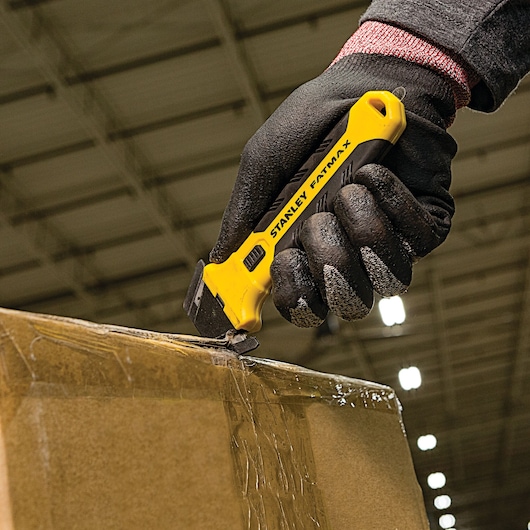Photo of STANLEY FMHT10361 utility knife cutting tape on a cardboard box, held by a gloved hand.
