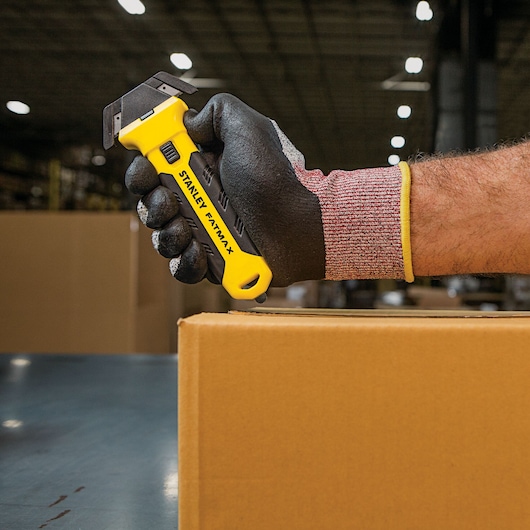 Photo of STANLEY FMHT10361 utility knife held in gloved hand, cutting a cardboard box in warehouse setting.