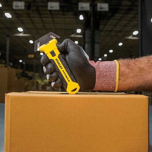 STANLEY FMHT10361 utility knife held by gloved hand cutting cardboard box in warehouse setting.