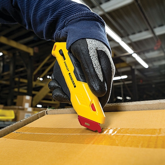 Stanley FMHT10362 yellow utility knife cutting box tape, work glove hand, warehouse background.