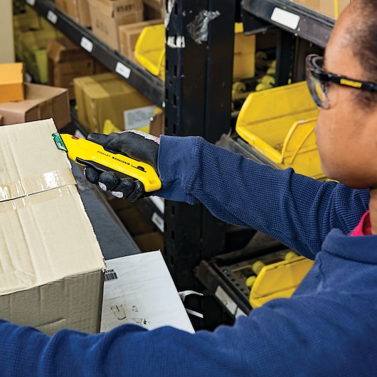 STANLEY FMHT10363 safety knife cutting cardboard box in warehouse, close-up of gloved hand holding tool.