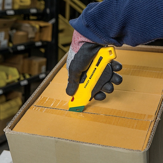 Stanley FMHT10363 yellow safety utility knife cutting box tape, close-up handheld in warehouse setting.