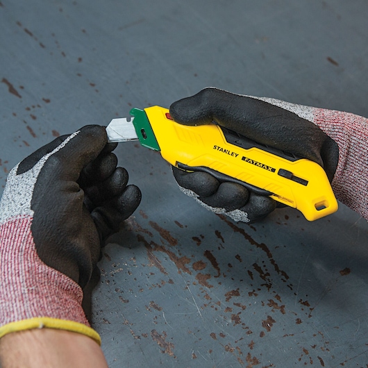 Stanley FMHT10363 yellow utility knife held in gloved hands on a worn metal surface.