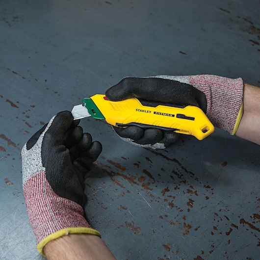 Photo of STANLEY FMHT10363 yellow utility knife held in gloved hands on a gray metal surface.