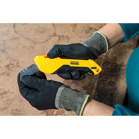 Stanley FMHT10369-0 utility knife with yellow and black handle, held by gloved hands, side view.
