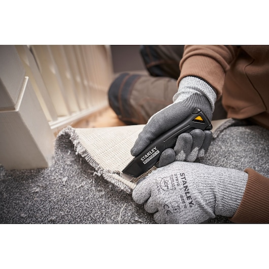 Photo of STANLEY FMHT10500-0 utility knife cutting carpet, gloved hands holding knife and carpet edge.