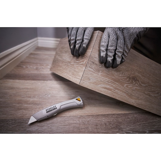 STANLEY FMHT10501-0 utility knife on wooden flooring beside gloved hands installing floor panel, angled view.