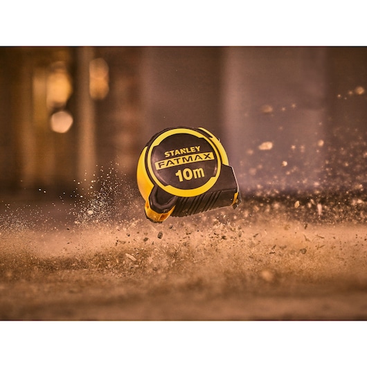 STANLEY FMHT33005-0 10m FATMAX tape measure in yellow and black, shown mid-air in dusty environment.