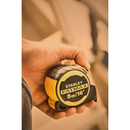 Photo of STANLEY FMHT33006-5 FatMax tape measure, yellow and black, held in hand, product close-up.