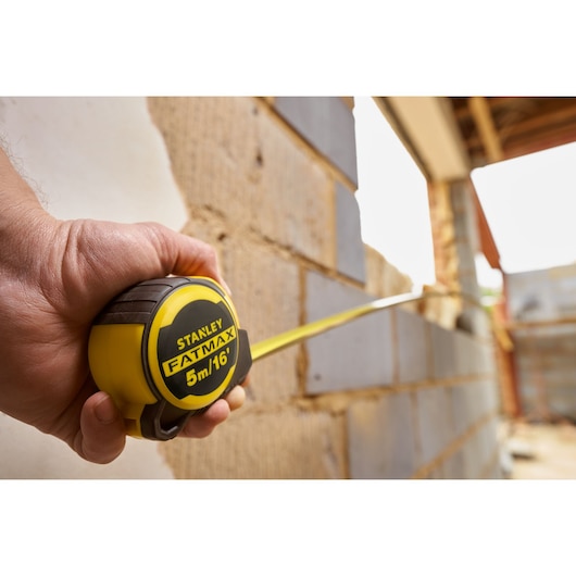 STANLEY FMHT33006-5 tape measure held, yellow and black, measuring wall, FATMAX logo visible.