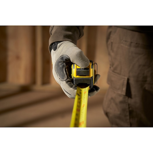 STANLEY FMHT33006-5 tape measure held by gloved hand, close-up, yellow and black tool in use.
