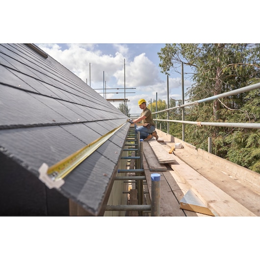 STANLEY FMHT38215-5 tape measure extended along roof edge, worker measuring on scaffolding, photography side angle.