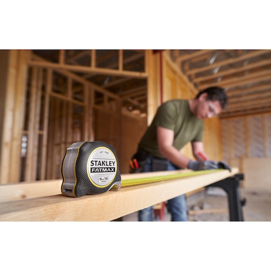 Photo of STANLEY FMHT38215-5 tape measure on wood in construction site, person measuring in background.