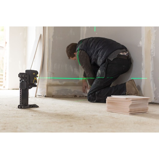 STANLEY FMHT77598-1 laser level projects green lines for wall alignment, worker kneeling nearby, stack of tiles visible.