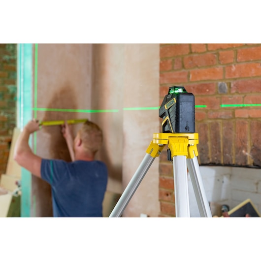 STANLEY FMHT77617-1 laser level on tripod projects green lines while person measures wall in room.