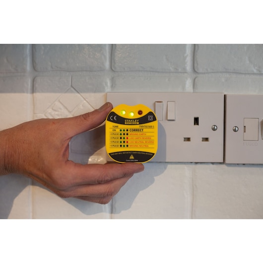 Photo of STANLEY FMHT82568-5 socket tester held near a wall outlet, showing indicator lights and yellow label.