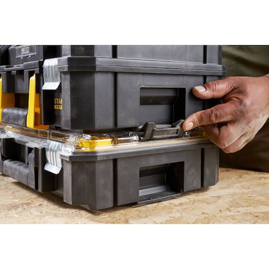 STANLEY FMST82967-9 tool box close-up, hand unlocking black and yellow stackable case on wood surface.