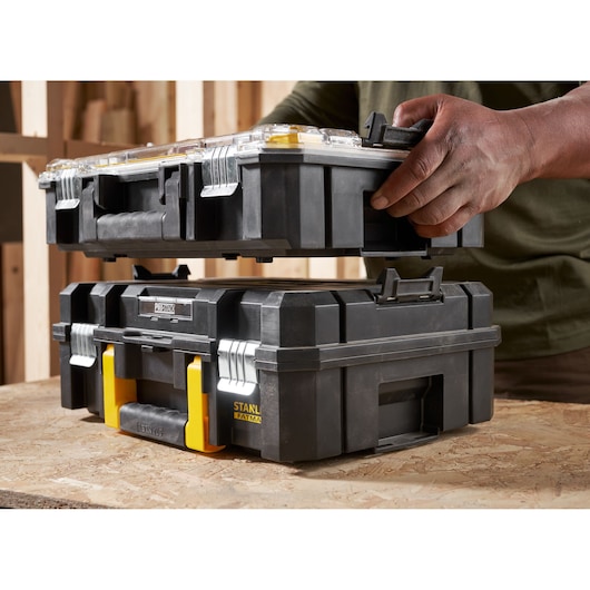 STANLEY FMST82967-9 stackable black toolbox with yellow accents, photographed on a workbench with a hand lifting the top box.