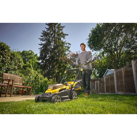 Photo of STANLEY SCMWP3401M1-XE lawn mower in use on green grass with wooden fence and bench in background.