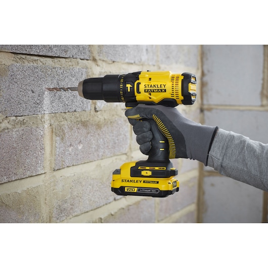 Stanley SFMCD711D21 cordless drill, yellow and black, in use drilling into brick wall, close-up side view.
