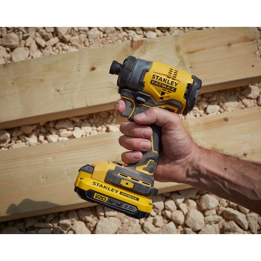 Photo of STANLEY SFMCF810D1K impact driver held in hand, yellow and black, on wood planks and stones.