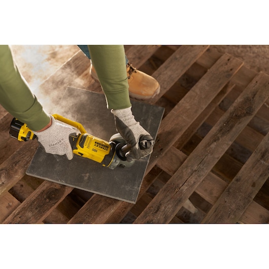 Photo of STANLEY SFMCG400M2K cordless angle grinder in use on a tile, held by gloved hands.