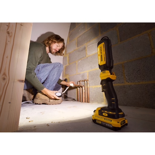 STANLEY SFMCL050B 18V FATMAX V20 Pivot Light on floor, illuminating pipes, person working in background.
