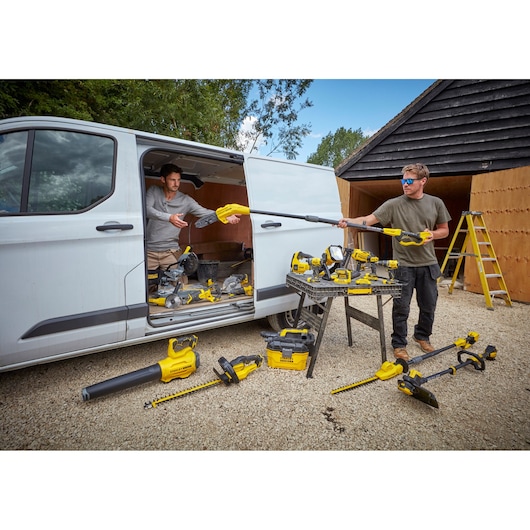 STANLEY SFMCPS620M1 cordless pole saw with yellow tools on ground and table near van and workshop, outdoor angle.