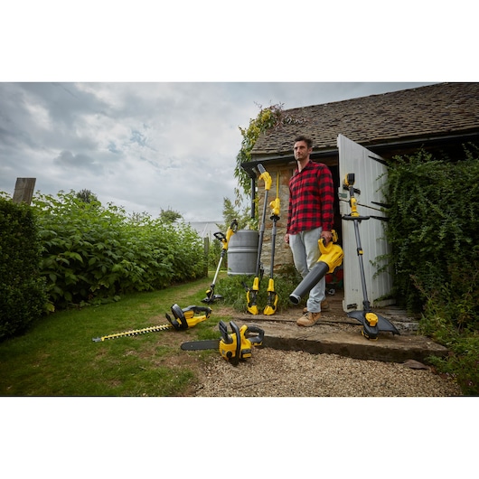 Photo of STANLEY SFMCPS620M1 garden power tools on ground near house, including chainsaw, hedge trimmer, and blower.