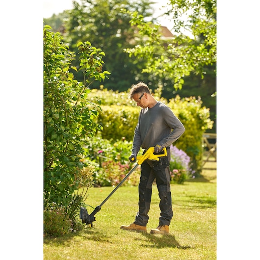 STANLEY SFMCST925M1 string trimmer in use outdoors, person trimming grass near bushes.