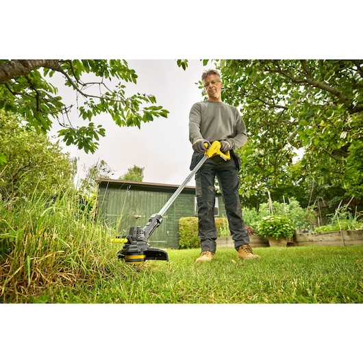 STANLEY SFMCST925M1 grass trimmer in use, person trimming lawn in garden with green shed in background.